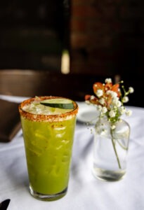 A green iced cocktail with a salted rim, garnished with a cucumber slice, sits beside a small vase of white flowers on a white tablecloth. © Design Thumbprint Marketing Agency