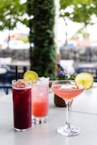 Three colorful cocktails with ice and lime slices on a white table outdoors, with blurred plants and patio seating in the background. © Design Thumbprint Marketing Agency